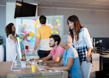 Un groupe de 3 personnes travaillant sur un ordi et deux autres personnes sont vers un tableau avec des post-it