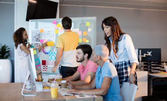 Un groupe de 3 personnes travaillant sur un ordi et deux autres personnes sont vers un tableau avec des post-it
