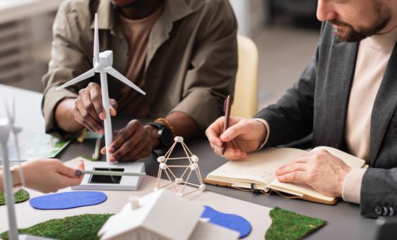 Des gens autour d'une table dont un qui écrit sur un cahier et un autre qui manipule une éolienne miniature